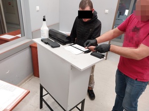 Zdjęcie przedstawia sylwetki dwóch osób, po prawej stronie zdjęcia widać ubranego w czerwoną koszulkę na krótki rękaw policjanta, który ma na dłoniach założone czarne rękawiczki ochronne i trzyma lewą dłoń zatrzymanego mężczyzny przyciskając ją do kartki ułożonej na jasnym stole. Twarz policjanta jest zasłonięta komputerowym filtrem. Zatrzymany mężczyzna jest ubrany w ciemny bezrękawnik i ciemną bluzę. Część twarzy zasłania mu czarny prostokąt.