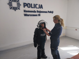 Zdjęcie przedstawia umundurowaną policjantkę, która stoi obok chłopca ubranego w policyjny strój wykorzystywany do zabezpieczenia meczów.