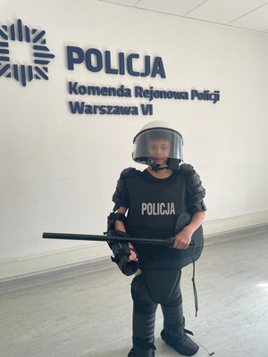 Zdjęcie przedstawia chłopca ubranego w policyjny strój wykorzystywany do zabezpieczenia meczów. W tle na ścianie widać granatowy napis: &quot;KOMENDA REJONOWA POLICJI WARSZAWA VI&quot;.