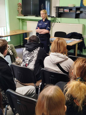 Zdjęcie przedstawia umundurowaną policjantkę, która stoi przed osobami siedzącymi na krzesłach.