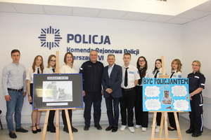 Zdjęcie przedstawia umundurowanego policjanta, dwóch mężczyzn, kobietę oraz kilkoro uczniów, którzy stoją w jednym rzędzie i uśmiechają się do zdjęcia.