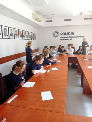 Zdjęcie przedstawia dwie umundurowane policjantki, które stoją na sali. Przy stole siedzą uczniowie, przed którymi leżą rozłożone białe kartki papieru.