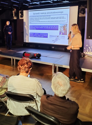 Zdjęcie przedstawia umundurowaną policjantkę oraz kobietę stojące na scenie po obu stronach ekranu na którym wyświetlany jest slajd prezentacji. Przed kobietami na krzesłach siedzą seniorzy.