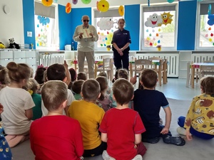 Zdjęcie przedstawia umundurowaną policjantkę i mężczyznę, którzy stoją przed siedzącymi na dywanie dziećmi.