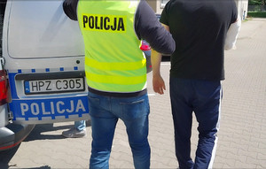 Zdjęcie przedstawia policjanta ubranego w czarną bluzkę na długi rękaw i seledynową odblaskową kamizelkę z napisem POLICJA na plecach, który prowadzi po swojej prawej stronie zatrzymanego mężczyznę. Po lewej stronie zdjęcia widać fragment oznakowanego radiowozu.