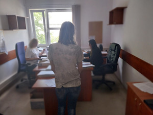 Zdjęcie przestawia kobietę, która stoi przed biurkiem przy którym siedzą dwie kobiety przed laptopami. Kobieta stoi tyłem do obiektywu aparatu. Twarze kobiet siedzących przy biurku są zasłonięte komputerowym filtrem.