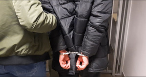 Zdjęcie przedstawia dwie sylwetki osób stojących tyłem do obiektywu aparatu przed okratowanymi drzwiami prowadzącymi do wyjścia. Osoba stojąca po prawej stronie zdjęcia ma kajdanki założone na rękach trzymanych z tyłu.