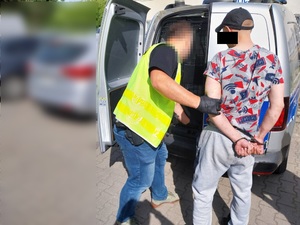 Zdjęcie prowadzi policjanta ubranego w odblaskową kamizelkę, który otwiera tylne drzwi do oznakowanego radiowozu. Po jego prawej stronie stoi zatrzymany mężczyzna. Jego twarz zasłania czarny prostokąt. Na rękach trzymanych z tyłu ma założone kajdanki.