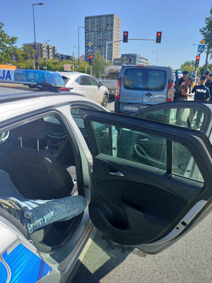 Zdjęcie przedstawia fragment sylwetki osoby siedzącej na tylnym siedzeniu w radiowozie. Przed radiowozem widać grupę umundurowanych policjantów stojących przy ciemnym samochodzie. W tle widać sygnalizację świetlną i inne stojące przed nią auta.