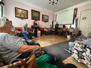 Zdjęcie przedstawia dwie policjantki, które stoją w pokoju wśród siedzących na krzesłach seniorów.