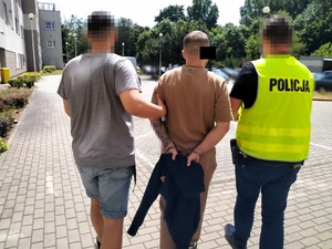 Zdjęcie przedstawia dwóch policjantów prowadzących pomiędzy sobą przez parking zatrzymanego mężczyznę. Mężczyzna ten jest ubrany w brązową koszulkę na krótki rękaw, część jego twarzy zasłania czarny prostokąt. Po jego prawej stronie idzie policjant ubrany w odblaskową kamizelkę z napisem POLICJA. Policjant idący po lewej stronie zatrzymanego jest ubrany w szarą koszulkę na krótki rękaw i niebieskie jeansowe spodenki. Twarze policjantów są zasłonięte.