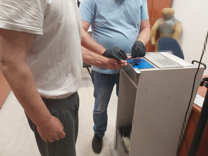 Zdjęcie przedstawia policjanta ubranego w niebieską koszulę na krótki rękaw, który ma na dłoniach czarne rękawiczki ochronne. Policjant trzyma w dłoniach dłoń mężczyzny i dociska jego palec do blatu biurka.