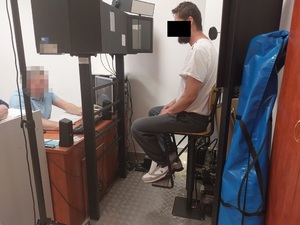Zdjęcie przedstawia policjanta w niebieskiej koszulce na krótki rękaw, który siedzi przed monitorem komputera. Jego twarz jest zasłonięta komputerowym filtrem. Przed nim na krześle, przed monitorami siedzi zatrzymany mężczyzna. Część jego twarzy zasłania czarny prostokąt.