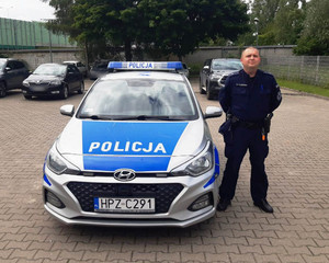 Zdjęcie przedstawia umundurowanego policjanta, który stoi przy zaparkowanym radiowozie.