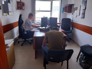 Zdjęcie przedstawia policjanta ubranego w jasną koszulkę na krótki rękaw, który siedzi przy biurku przed otwartym laptopem. Tyłem do zdjęcia siedzi zatrzymany mężczyzna. Ubrany jest w brązową koszulkę na krótki rękaw i długie spodnie jeansowe.
