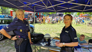 Zdjęcie przedstawia dwie umundurowane policjantki, które stoją przy stolikach z gadżetami i uśmiechają się do zdjęcia.