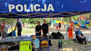 Zdjęcie przedstawia dwie umundurowane policjantki, które stoją przy stolikach z gadżetami pod namiotem z napisem POLICJA i uśmiechają się do zdjęcia.