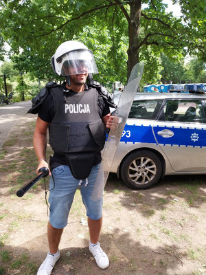 Zdjęcie przedstawia mężczyznę ubranego w sprzęt policyjny w postaci kasku i kamizelki kuloodpornej. W ręku trzyma czarną pałkę szturmową.