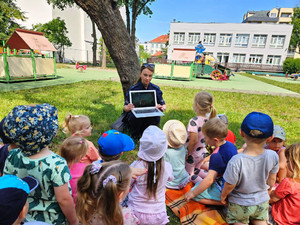 Zdjęcie przedstawia policjantkę, która prezentuje na laptopie film edukacyjny dzieciom siedzącym na kocu.