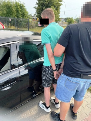 Zdjęcie przedstawia policjanta ubranego w ciemną koszulkę na krótki rękaw i niebieskie spodenki jeansowe, który stoi za zatrzymanym mężczyzną. Twarz policjanta jest zasłonięta komputerowym filtrem. Mężczyzna jest ubrany w niebieską koszulkę na krótki rękaw, ciemne krótkie spodenki i jasne sportowe buty. Na rekach trzymanych z tyłu ma założone kajdanki. Cześć jego twarzy zasłania czarny prostokąt. Obaj stoją tyłem do obiektywu aparatu przy ciemnym samochodzie.