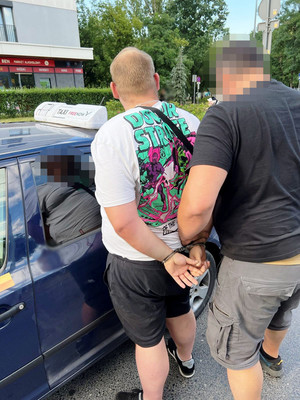 Zdjęcie przedstawia policjanta ubranego w ciemną koszulkę na krótki rękaw i niebieskie spodenki, który przytrzymuje za ręce stojącego przed nim zatrzymanego mężczyznę. Mężczyzna ten ubrany jest w białą koszulkę na krótki rękaw z kolorowymi rysunkami na plecach i ciemne krótki spodenki. Mężczyzna ten ma założone kajdanki  na rękach trzymanych z tyłu. Twarz policjanta jest zasłonięta komputerowym filtrem. Obaj stoją przy zaparkowanym ciemnoniebieskim samochodzie.