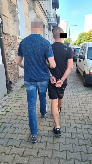 Zdjęcie przedstawia policjanta ubranego w granatowa koszulkę na krótki rękaw, który prowadzi po chodniku, po swojej prawej stronie zatrzymanego mężczyznę. Mężczyzna ten ubrany jest w ciemną koszulkę na krótki rękaw i ciemne spodenki. Na rekach trzymanych z tyłu ma założone kajdanki.