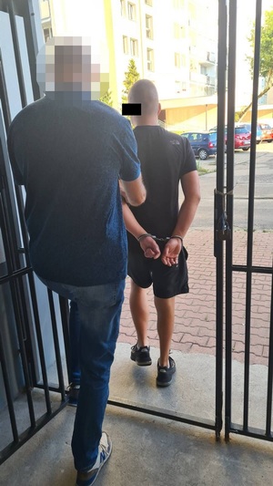 Zdjęcie przedstawia policjanta ubranego w granatowa koszulkę na krótki rękaw, który prowadzi po chodniku, po swojej prawej stronie zatrzymanego mężczyznę. Mężczyzna ten ubrany jest w ciemną koszulkę na krótki rękaw i ciemne spodenki. Na rekach trzymanych z tyłu ma założone kajdanki.