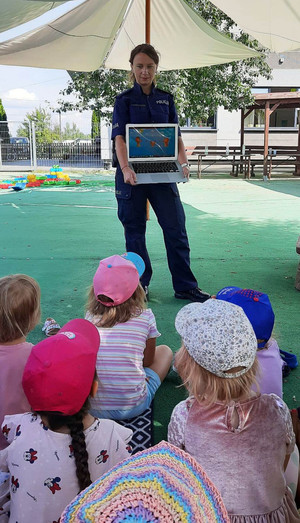 Zdjęcie przedstawia policjantkę stojącą przed dziećmi siedzącymi na ziemi. Policjantka trzyma w dłoniach laptop, na którym wyświetla dzieciom film edukacyjny.