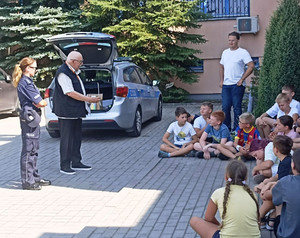 Zdjęcie przedstawia policjantkę w mundurze i mężczyznę stojących przed siedzącymi na chodniku uczniami.