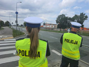 Zdjęcie przedstawia sylwetki policjanta i policjantki ubranych w kamizelki odblaskowe i stojących tyłem na zdjęciu.