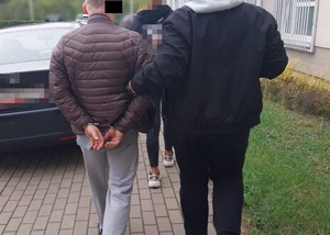 Zdjęcie przedstawia ubranego na ciemno policjanta, który prowadzi po swojej lewej stronie zatrzymanego mężczyznę.