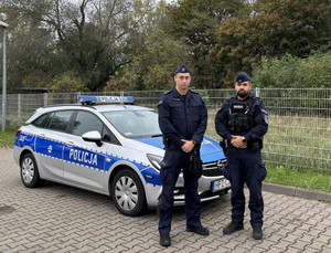 Zdjęcie przedstawia dwóch umundurowanych policjantów, którzy stoją przed oznakowanym radiowozem.