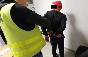 Zdjęcie przedstawia policjanta ubranego w odblaskową kamizelkę, który zakłada kajdanki zatrzymanemu mężczyźnie na ręce trzymane z tyłu. Obaj znajdują się w pomieszczeniu dla osób zatrzymanych. Zatrzymany mężczyzna ma na głowie czerwoną czapkę z daszkiem. Część jego twarzy zasłania czarny prostokąt.