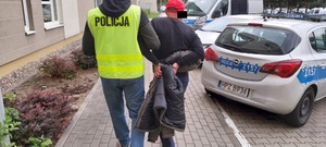 Zdjęcie przedstawia policjanta ubranego w odblaskową kamizelkę, który prowadzi po dziedzińcu po swojej prawej stronie zatrzymanego mężczyznę. Mężczyzna ten ma kajdanki założone na rękach trzymanych z tyłu. Ma na głowie czerwoną czapkę z daszkiem. Część jego twarzy zasłania czarny prostokąt. W tle widać zaparkowane oznakowane radiowozy.