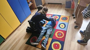 Zdjęcie przedstawia umundurowaną policjantkę, która pochyla się nad dzieckiem leżącym na dywanie. Obok niej widać inne kucające na dywanie dziecko.