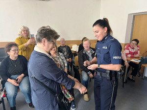 Zdjęcie przedstawia umundurowaną policjantkę i siedzących na krześle seniorów. Obok niej stoi kobieta, która ma na rękach założone kajdanki.