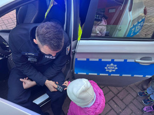 Zdjęcie przedstawia umundurowanego policjanta siedzącego w radiowozie, który pokazuje małemu dziecku pilota od sygnałów dźwiękowych i świetlnych.