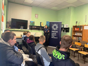 Zdjęcie przedstawia policjantkę stojącą przed młodzieżą siedzącą na krzesłach w sali. Policjantka stoi obok plakatu z napisem KOMENDA REJONOWA POLICJI WARSZAWA VI.