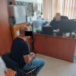 Zdjęcie przedstawia sylwetkę policjanta ubranego na ciemno, który siedzi za biurkiem przed monitorem laptopa. Na krześle przodem do niego siedzi zatrzymany mężczyzna. Mężczyzna ten jest ubrany w ciemną koszulkę na krótki rękaw i niebieskie spodnie jeansowe. Na rękach ma  założone kajdanki.