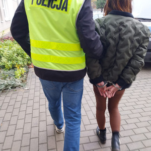 Zdjęcie przedstawia policjanta ubranego w seledynową kamizelkę, który prowadzi po swojej prawej stronie zatrzymaną kobietę. Kobieta ta ma kajdanki na rękach.