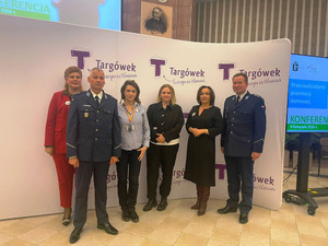 Zdjęcie przedstawia policjanta stojącego z innymi osobami na tle plakatu.