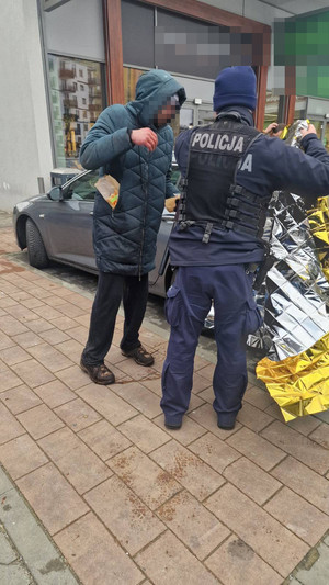 Zdjęcie przedstawia policjanta, który  ma w dłoniach srebrno-złotą folię ochronną, obok niego stoi mężczyzna z kapturem na głowie.