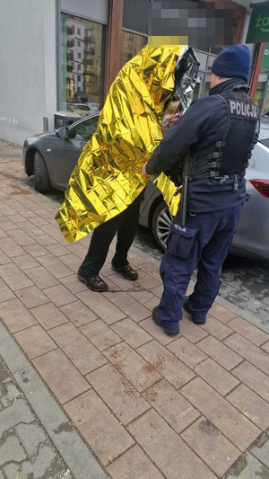 Zdjęcie przedstawia policjanta, który okrywa srebrno-złotą folią ochronną stojącego obok niego mężczyznę.