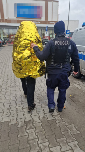 Zdjęcie przedstawia policjanta, który prowadzi po swojej lewej stronie mężczyznę okrytego srebrno-złotą folią ochronną.