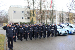 Zdjęcie przedstawia umundurowanych policjantów, który stoją w szeregu obok zaparkowanych radiowozów, które mają włączone światła błyskowe.