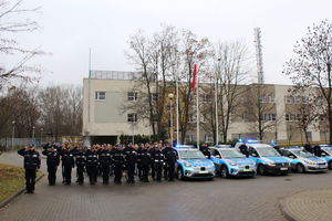 Zdjęcie przedstawia umundurowanych policjantów, który stoją w szeregu obok zaparkowanych radiowozów, które mają włączone światła błyskowe.