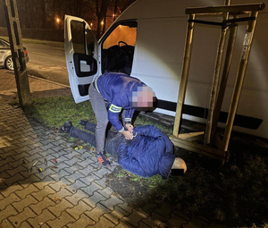 Zdjęcie przedstawia policjanta ubranego w niebieską kurtkę, który pochyla się nad mężczyzną leżącym na ziemi. Za nim widać zaparkowanego busa. Mężczyzna leżący na ziemi trzyma ręce na plecach.