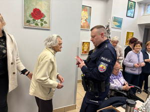 Zdjęcie przedstawia policjanta stojącego przed starszą kobietę i trzymającego w dłoniach ulotki profilaktyczne.