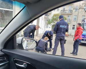 Zdjęcie przedstawia trzech policjantów pochylających się nad leżącym na ziemi mężczyzną i jednego stojącego nad nimi. Obok niego widać osobę stojącą w czerwonej kurtce. Jej twarz jest zasłonięta.