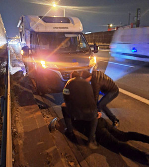 Zdjęcie przedstawia dwóch ubranych na ciemno policjantów, którzy pochylają się nad leżącym na ziemi ubranym na ciemno mężczyzną. Przed nimi widać zaparkowany biały pojazd.